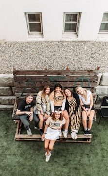 A group of six people are sitting and standing on a wooden bench outdoors, smiling and appearing relaxed. They are casually dressed, with some wearing Purdue University apparel. Behind them, there is a rustic wooden panel with Greek letters, and the scene is set near a building with two small windows. The area appears to be a patio or yard with a green artificial grass surface.