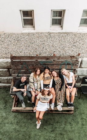 A group of six people are sitting and standing on a wooden bench outdoors, smiling and appearing relaxed. They are casually dressed, with some wearing Purdue University apparel. Behind them, there is a rustic wooden panel with Greek letters, and the scene is set near a building with two small windows. The area appears to be a patio or yard with a green artificial grass surface.
