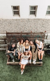 A group of six people are sitting and standing on a wooden bench outdoors, smiling and appearing relaxed. They are casually dressed, with some wearing Purdue University apparel. Behind them, there is a rustic wooden panel with Greek letters, and the scene is set near a building with two small windows. The area appears to be a patio or yard with a green artificial grass surface.