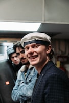 Three young men appear together indoors, with the one in the foreground smiling widely. He is wearing a white beret and a blue checkered blazer. The other two are positioned slightly behind, wearing casual jackets and hats. The scene is dimly lit, with a modern, urban vibe.