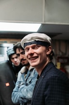 Three young men appear together indoors, with the one in the foreground smiling widely. He is wearing a white beret and a blue checkered blazer. The other two are positioned slightly behind, wearing casual jackets and hats. The scene is dimly lit, with a modern, urban vibe.