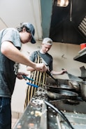 Two individuals are cooking in a kitchen, focused on their tasks. One is wearing a striped apron and the other a casual T-shirt. They are working with various cooking utensils and pans on a stovetop. The kitchen shows signs of active use, with food remnants visible.