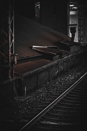 The image features a dimly lit industrial setting, dominated by a dark, angled metal roof structure and adjacent train tracks. The texture of the roof is highlighted by the sparse lighting, creating shadows and contrast. A part of a metal support beam and signs of industrial warehousing are visible, adding to the gritty and utilitarian atmosphere.