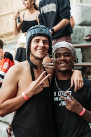 Two young men are posing happily for the camera. One is shirtless, wearing an apron and helmet, and flashing a peace sign with both hands. The other is dressed in a black shirt with a headscarf, also making a peace sign with one hand. They have their arms around each other. In the background, there are other people, including a girl in a black tank top and a man wearing a dark sweatshirt.