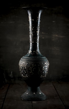 A close-up of a handcrafted metal vase with a textured green patina finish, resting on a beige surface.