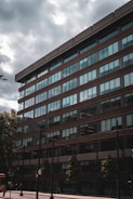 A multi-story office building with large glass windows and brown exterior panels is seen from a low angle perspective. A cloudy sky looms overhead, and there are trees and street signs in the foreground. The area appears to be urban with some pedestrians visible near the bottom of the building.