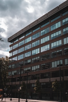 A multi-story office building with large glass windows and brown exterior panels is seen from a low angle perspective. A cloudy sky looms overhead, and there are trees and street signs in the foreground. The area appears to be urban with some pedestrians visible near the bottom of the building.