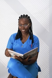 A person in blue scrubs is seated on a stool while holding an open book and smiling. The background has a pattern of diagonal shadows, and they are wearing glasses, a necklace, and have braided hair. There is a pen visible in the breast pocket of the scrubs.