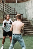 Two individuals are engaged in a boxing match. One is wearing boxing gloves and a white hoodie, facing the shirtless opponent with tattoos. They stand outdoors on green artificial turf by a set of concrete stairs, fenced by metal railings.