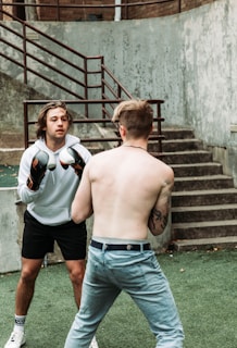 Two individuals are engaged in a boxing match. One is wearing boxing gloves and a white hoodie, facing the shirtless opponent with tattoos. They stand outdoors on green artificial turf by a set of concrete stairs, fenced by metal railings.