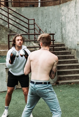 Two individuals are engaged in a boxing match. One is wearing boxing gloves and a white hoodie, facing the shirtless opponent with tattoos. They stand outdoors on green artificial turf by a set of concrete stairs, fenced by metal railings.