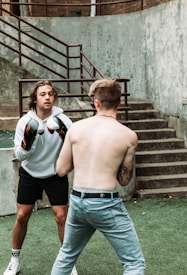 Two individuals are engaged in a boxing match. One is wearing boxing gloves and a white hoodie, facing the shirtless opponent with tattoos. They stand outdoors on green artificial turf by a set of concrete stairs, fenced by metal railings.