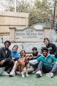 Seven young men and a dog are gathered in front of a sign that reads 'Delta Sigma Phi ΔΣΦ Chapter, 1899 - 1957'. They are sitting and kneeling on a lawn with a wooden fence and stairs in the background.