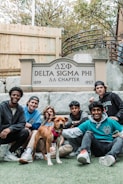Seven young men and a dog are gathered in front of a sign that reads 'Delta Sigma Phi ΔΣΦ Chapter, 1899 - 1957'. They are sitting and kneeling on a lawn with a wooden fence and stairs in the background.