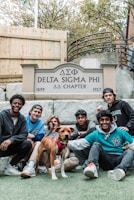 Seven young men and a dog are gathered in front of a sign that reads 'Delta Sigma Phi ΔΣΦ Chapter, 1899 - 1957'. They are sitting and kneeling on a lawn with a wooden fence and stairs in the background.