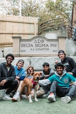 Seven young men and a dog are gathered in front of a sign that reads 'Delta Sigma Phi ΔΣΦ Chapter, 1899 - 1957'. They are sitting and kneeling on a lawn with a wooden fence and stairs in the background.