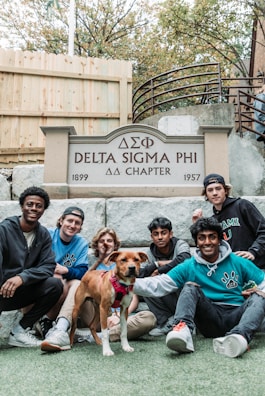 Seven young men and a dog are gathered in front of a sign that reads 'Delta Sigma Phi ΔΣΦ Chapter, 1899 - 1957'. They are sitting and kneeling on a lawn with a wooden fence and stairs in the background.