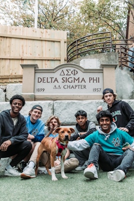 Seven young men and a dog are gathered in front of a sign that reads 'Delta Sigma Phi ΔΣΦ Chapter, 1899 - 1957'. They are sitting and kneeling on a lawn with a wooden fence and stairs in the background.