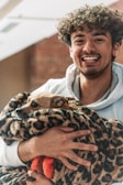 A person with curly hair is smiling brightly while holding a small dog wrapped in a leopard-print blanket. The dog looks content and is also wrapped partially in a red and orange knit fabric. The background features a brick wall and a soft indoor setting.