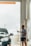 A person wearing shorts and a T-shirt is holding a water hose, washing a black car in a car wash bay. The scene is indoors, with light streaming in, and there are signs of water droplets on the vehicle.