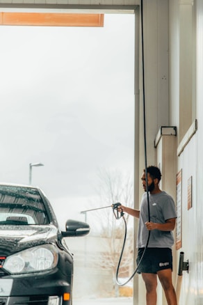 A person wearing shorts and a T-shirt is holding a water hose, washing a black car in a car wash bay. The scene is indoors, with light streaming in, and there are signs of water droplets on the vehicle.