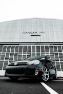 A black Volkswagen car is parked in front of a large hangar with 'HANGAR NO.1' written on it. The hangar features large, industrial-style doors made of metal, creating a stark, imposing backdrop. The car is positioned at an angle, emphasizing its sporty design and glossy finish.