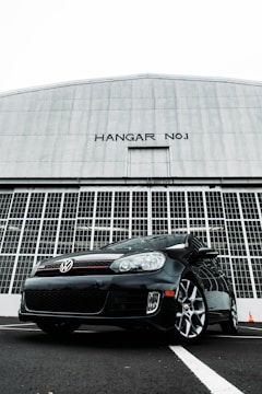 A black Volkswagen car is parked in front of a large hangar with 'HANGAR NO.1' written on it. The hangar features large, industrial-style doors made of metal, creating a stark, imposing backdrop. The car is positioned at an angle, emphasizing its sporty design and glossy finish.