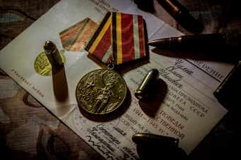 A commemorative medal with '1945-1975' and a figure holding a banner is placed on top of official-looking documents. The background features bullets and a medal ribbon with alternating yellow and red stripes, suggesting a military theme.