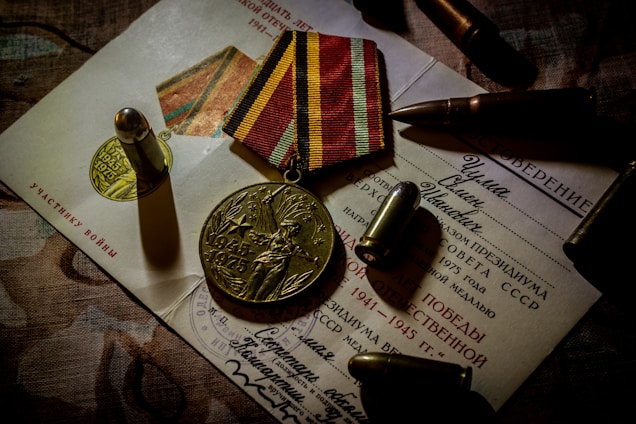 A commemorative medal with '1945-1975' and a figure holding a banner is placed on top of official-looking documents. The background features bullets and a medal ribbon with alternating yellow and red stripes, suggesting a military theme.
