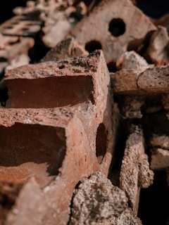 A close-up view of several reddish-brown bricks stacked together, with rough and uneven surfaces. Some of the bricks have holes, adding to the texture of the composition. The lighting casts shadows and highlights the coarse texture of the material.