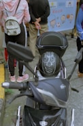 A black motorcycle with a helmet that has a detailed design resembling a face with teeth is parked on the street. Behind the motorcycle, a person wearing a pink and white hoodie and carrying a patterned backpack stands with others. The background features a banner with cartoon characters and text.