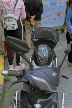 A black motorcycle with a helmet that has a detailed design resembling a face with teeth is parked on the street. Behind the motorcycle, a person wearing a pink and white hoodie and carrying a patterned backpack stands with others. The background features a banner with cartoon characters and text.