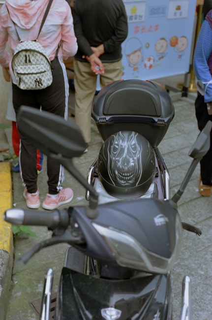 A black motorcycle with a helmet that has a detailed design resembling a face with teeth is parked on the street. Behind the motorcycle, a person wearing a pink and white hoodie and carrying a patterned backpack stands with others. The background features a banner with cartoon characters and text.
