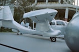 A silver aircraft with a sleek design is parked on a paved surface next to a small silver car. There is a large spherical object nearby and a person walking past the scene. The background includes trees and the structure of a building under construction.