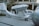 A silver aircraft with a sleek design is parked on a paved surface next to a small silver car. There is a large spherical object nearby and a person walking past the scene. The background includes trees and the structure of a building under construction.
