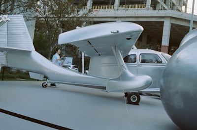 A silver aircraft with a sleek design is parked on a paved surface next to a small silver car. There is a large spherical object nearby and a person walking past the scene. The background includes trees and the structure of a building under construction.