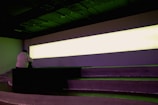 A person sits on tiered seating in a dimly lit room with a large, horizontally elongated, illuminated light panel dominating the wall. The atmosphere suggests a contemplative setting.