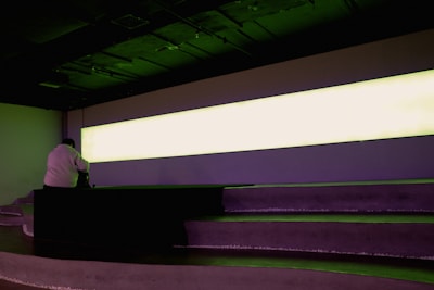 A person sits on tiered seating in a dimly lit room with a large, horizontally elongated, illuminated light panel dominating the wall. The atmosphere suggests a contemplative setting.