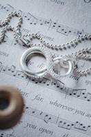 A silver chain necklace with intertwined rings rests on a sheet of printed music. The rings have an intricate design, and the sheet music contains both musical notes and lyrics. The image also includes a blurred element in the foreground.