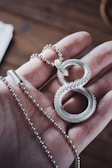 A hand holding a silver necklace with a decorative pendant resembling an infinity symbol. The pendant is detailed with small circular patterns, and the necklace chain consists of small, round beads. The background features a blurred, wooden surface.