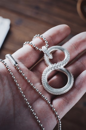 A hand holding a silver necklace with a decorative pendant resembling an infinity symbol. The pendant is detailed with small circular patterns, and the necklace chain consists of small, round beads. The background features a blurred, wooden surface.