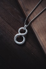 A silver necklace with a pendant shaped like an infinity symbol is placed diagonally across a wooden surface. The chain is composed of small interconnected links and the pendant features a beaded texture, adding an intricate detail to its design. The lighting casts subtle shadows, enhancing the elegance of the piece.