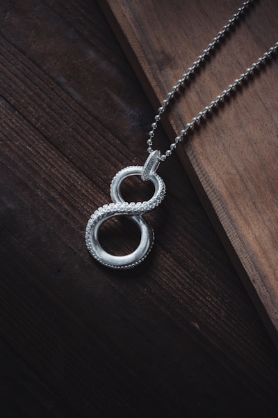 A silver necklace with a pendant shaped like an infinity symbol is placed diagonally across a wooden surface. The chain is composed of small interconnected links and the pendant features a beaded texture, adding an intricate detail to its design. The lighting casts subtle shadows, enhancing the elegance of the piece.