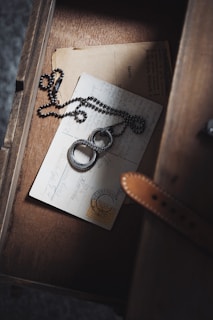 A vintage postcard with handwriting and a stamp lies in a wooden drawer. A round, metallic necklace with intricate designs is placed on top, wrapped in a casual manner. The drawer has a warm, rustic appearance, adding to the nostalgic feel of the scene.