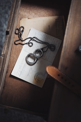 A vintage postcard with handwriting and a stamp lies in a wooden drawer. A round, metallic necklace with intricate designs is placed on top, wrapped in a casual manner. The drawer has a warm, rustic appearance, adding to the nostalgic feel of the scene.