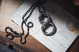 A necklace with a black chain and pendant shaped like two interlocked rings is placed over vintage postcards and letters. The surface is wooden, adding a rustic touch.