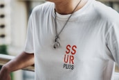 A person is wearing a white t-shirt with the text 'SSUR PLUS' in red and black letters on the front. The individual is also wearing a necklace with a chain and two round pendants. The background appears to have a railing and an out-of-focus cityscape or buildings.