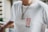A person is wearing a white t-shirt with the text 'SSUR PLUS' in red and black letters on the front. The individual is also wearing a necklace with a chain and two round pendants. The background appears to have a railing and an out-of-focus cityscape or buildings.