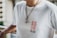 A person is wearing a white t-shirt with the text 'SSUR PLUS' in red and black letters on the front. The individual is also wearing a necklace with a chain and two round pendants. The background appears to have a railing and an out-of-focus cityscape or buildings.