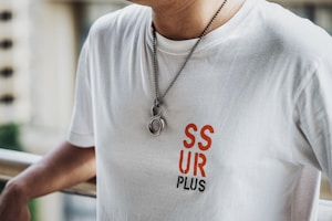 A person is wearing a white t-shirt with the text 'SSUR PLUS' in red and black letters on the front. The individual is also wearing a necklace with a chain and two round pendants. The background appears to have a railing and an out-of-focus cityscape or buildings.